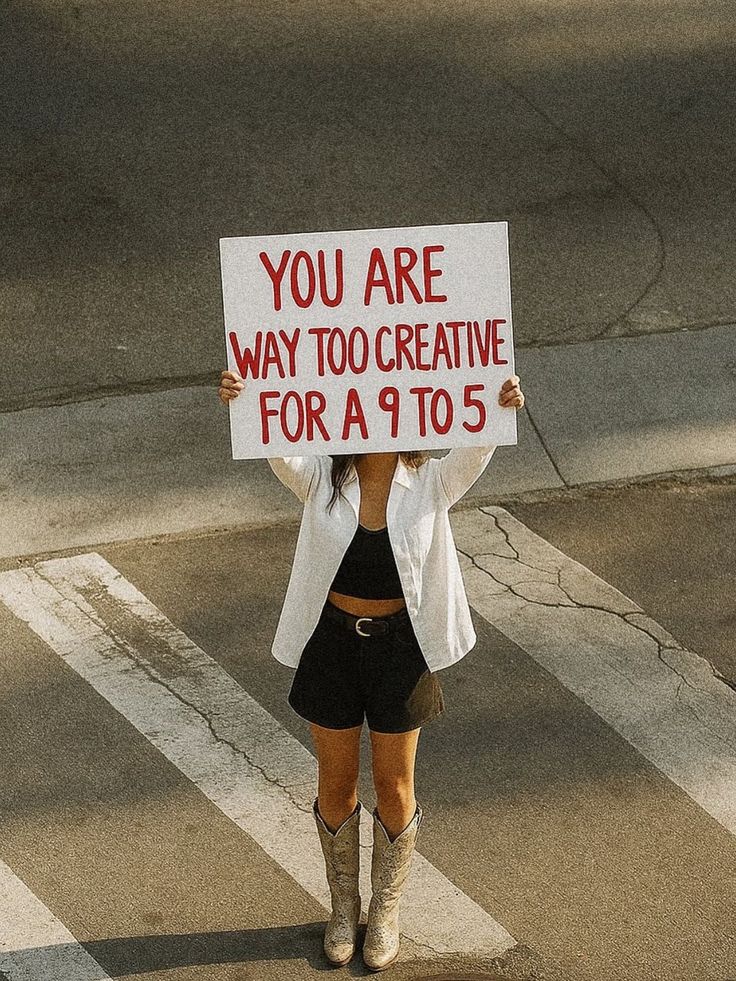 A woman on a crosswalk holding a sign that reads You Are Way Too Creative For A 9 To 5